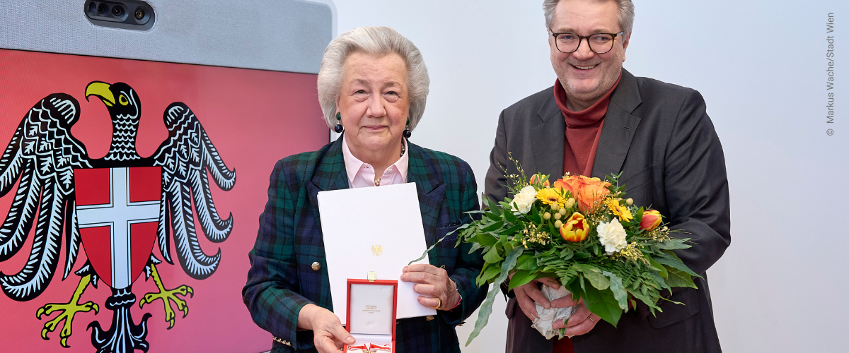 Christl Weinberger mit Auszeichnung in der Hand neben Stadtrat Peter Hacker mit Blumen in der Hand vor Screen mit Wien Wappen