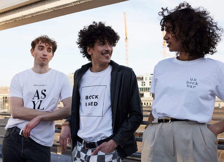 Drei junge Menschen stehen zusammen und tragen Shirts mit Botschaften wie "unbockbar", "Bock i ned" und "Menschenrecht Asyl" aus dem Bock Shop