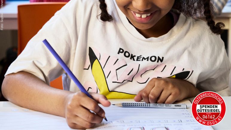 Ein Mädchen mit einem Pokemon-Shirt sitzt bei einem Tisch und schreibt ihre Hausünung.
