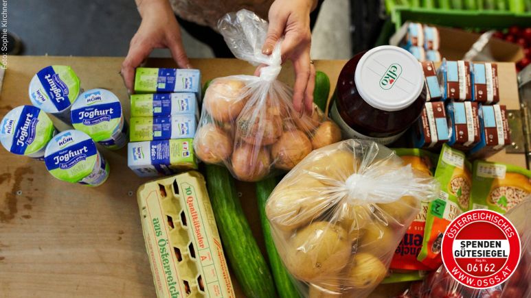 Zwei Hände, die gerade verschieden Lebensmittel wie Butter, Eier, Kartoffeln, Gurken auf einem Tisch herrichten.