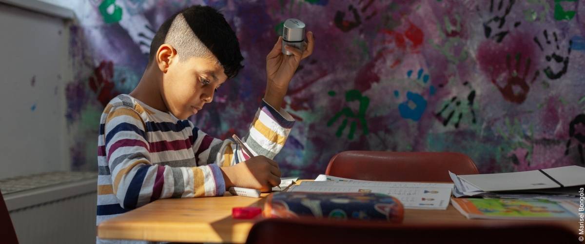 Ein Bub sitzt im Ute Bock Bildunszentrum an einem Tisch und schreibt seine Hausübung. Er hält einen Spitzer in der Hand und trägt ein bunt gestrieftes Shirt.