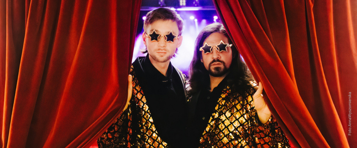 Die Magier Siegfried & Joy stehen in Glitzeranzügen in der Mitte eines roten Vorhangs und tragen goldene Sonnenbrillen