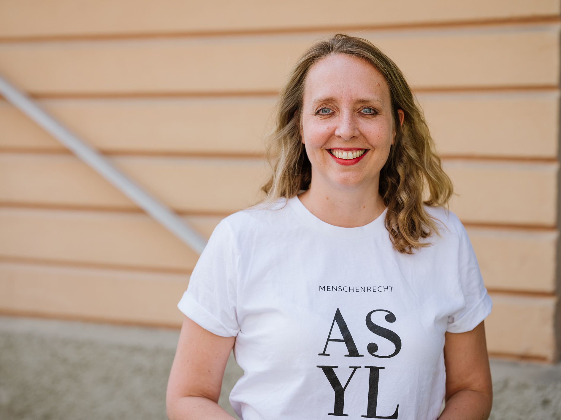eine Frau mit weißen Shirt mit dem Aufdrucj ASYL steht vor einer orangfarbenen Hausmauer