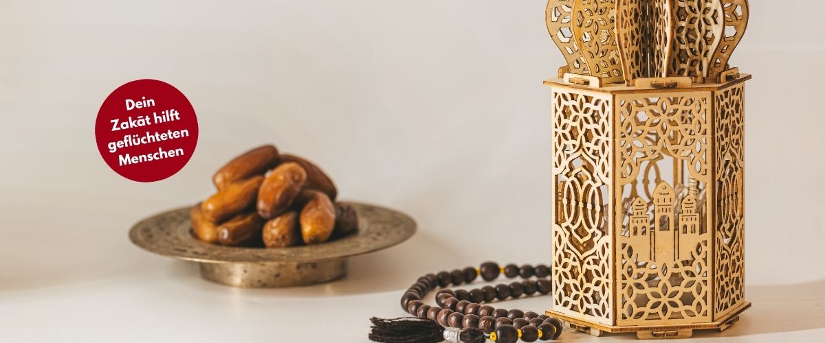 Datteln auf einem goldenen Teller, eine Ramadanlaterne aus Holz daneben. Davor eine rote Bubble mit dem Text "Dein Zakat hilft geflüchteten Menschen"