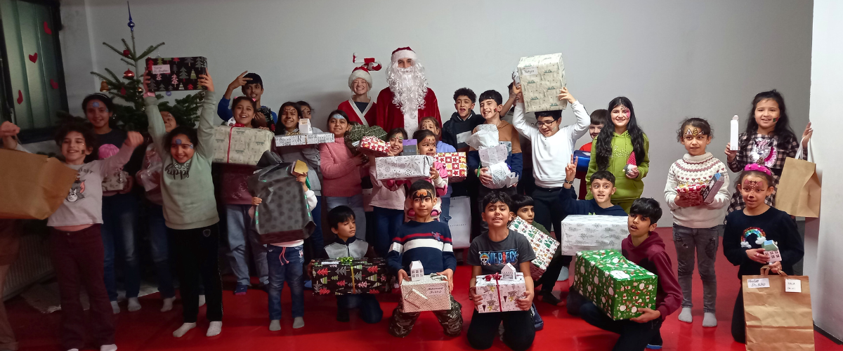Viele Kinder mit bunten Geschenken, ind er Mitte stehen der Weihnachtsmann und seine Elfe