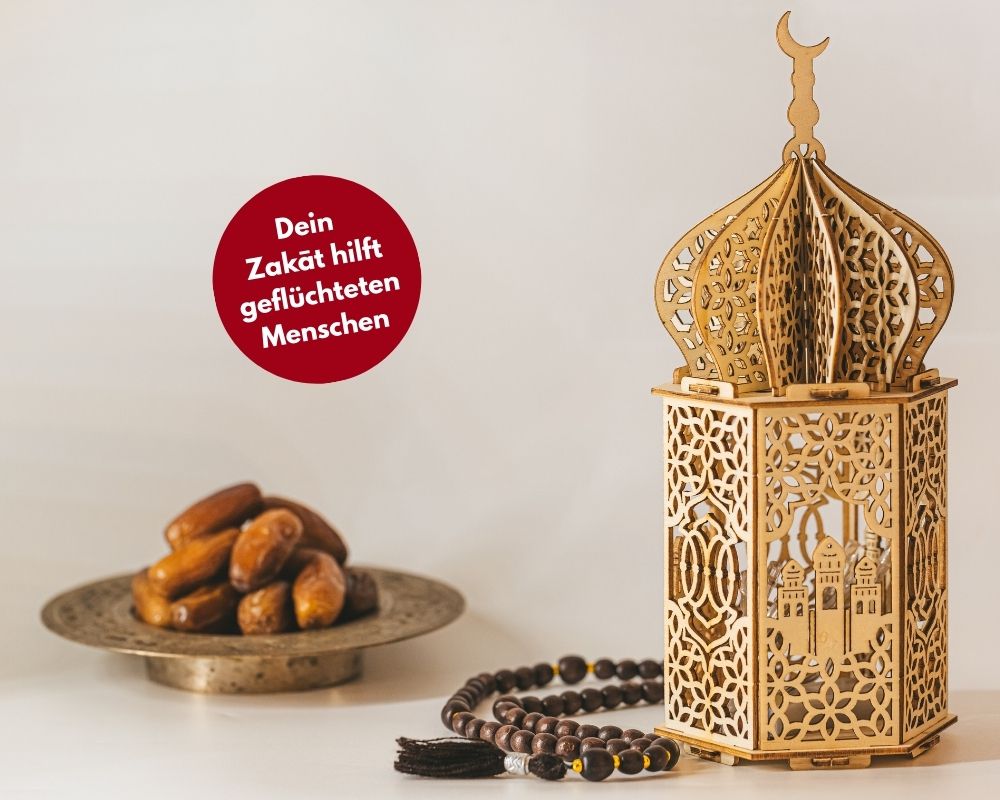 Datteln auf einem goldenen Teller, eine Ramadanlaterne aus Holz daneben. Davor eine rote Bubble mit dem Text "Dein Zakat hilft geflüchteten Menschen"