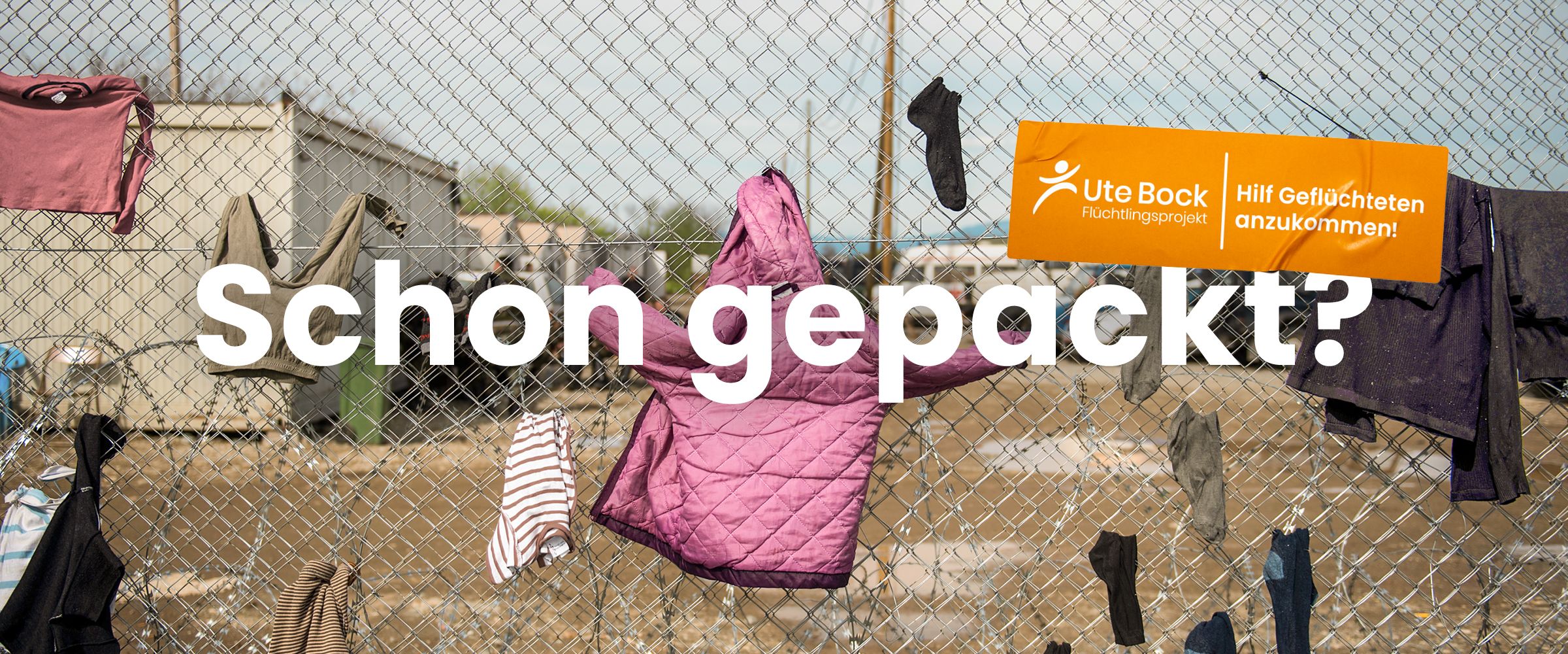 Schriftzug "Schon gepackt?" steht über Kinderkleidung, die am Zaun eines Flüchtlingsheims hängt