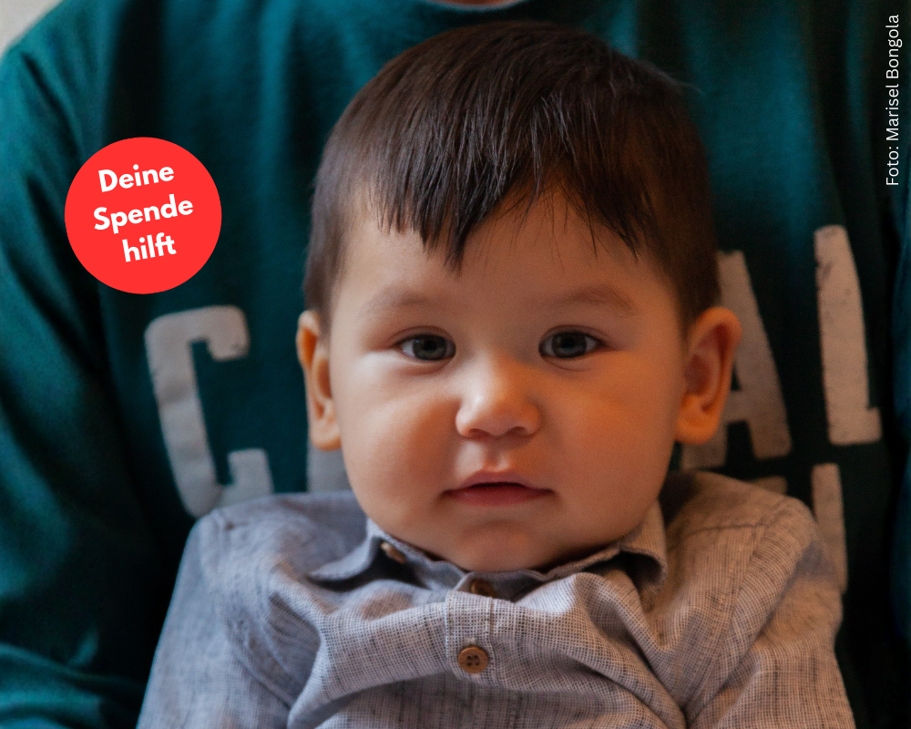 Kleinkind sitzt auf dem Schoß seines Papas. Das Kind schaut direkt in die Kamera. Daneben eine rote Bubble "Deine Spende hilft"