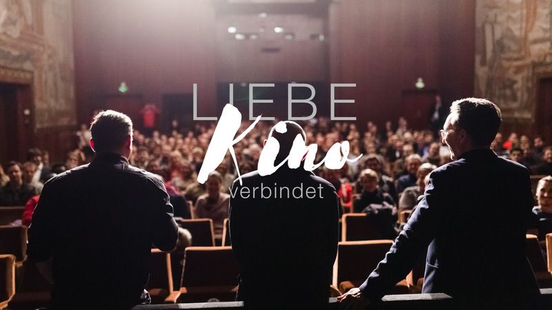 3 Menschen stehen vor einem Kinopublikum. Slogan "Kinoliebe verbindet"