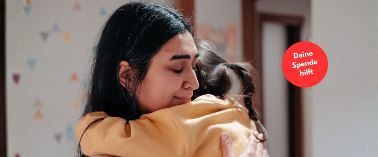 Eine Frau mit dunklen Haaren umarmt ein Kleinkind. Sie sieht erleichtert aus.