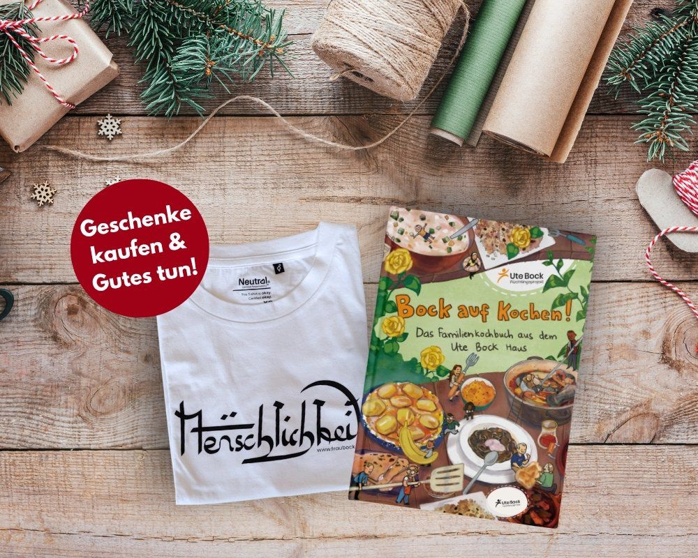 Ein Holztisch, darauf liegen weihnachtliches Verpackungsmaterial, ein weißes Shirt mit dem Aufdruck "Menschlichkeit" und ein Kochbuch "Bock auf Kochen". Weiter oben eine rote Bubble "Geschenke kaufen & Gutes tun"