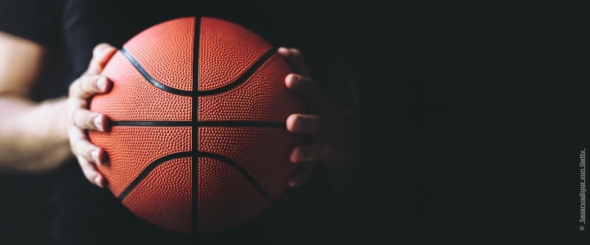 Schwarzer Hintergrund, im Vordergrund zwei Hände, die einen Basketball halten